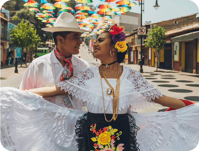A smiling couple in traditional Mexican clothing dances on a colorful street decorated with hanging umbrellas.